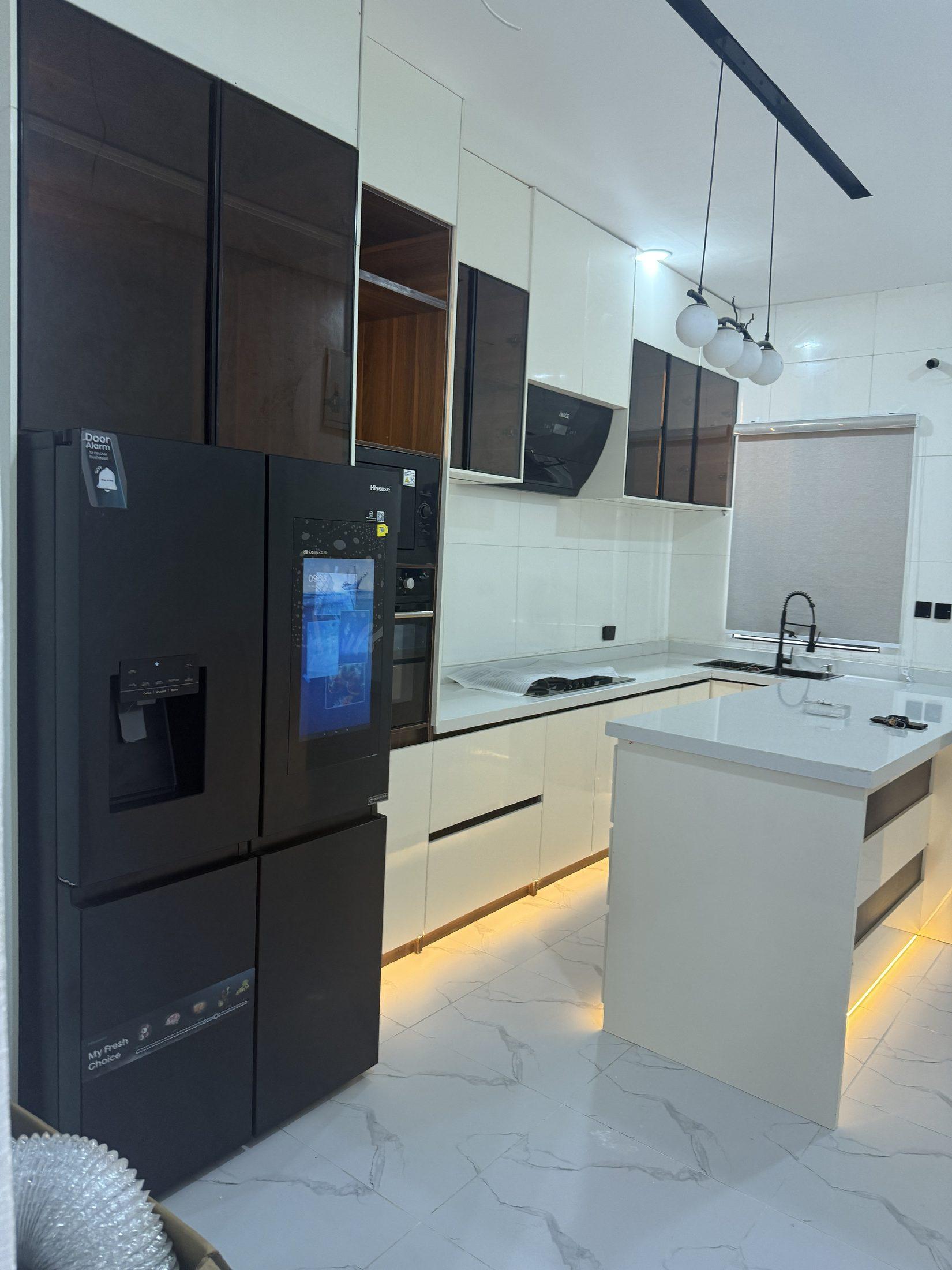 Kitchen island with a white stone top and warm plinth lighting, set against cream gloss cabinetry and smoked glass wall units