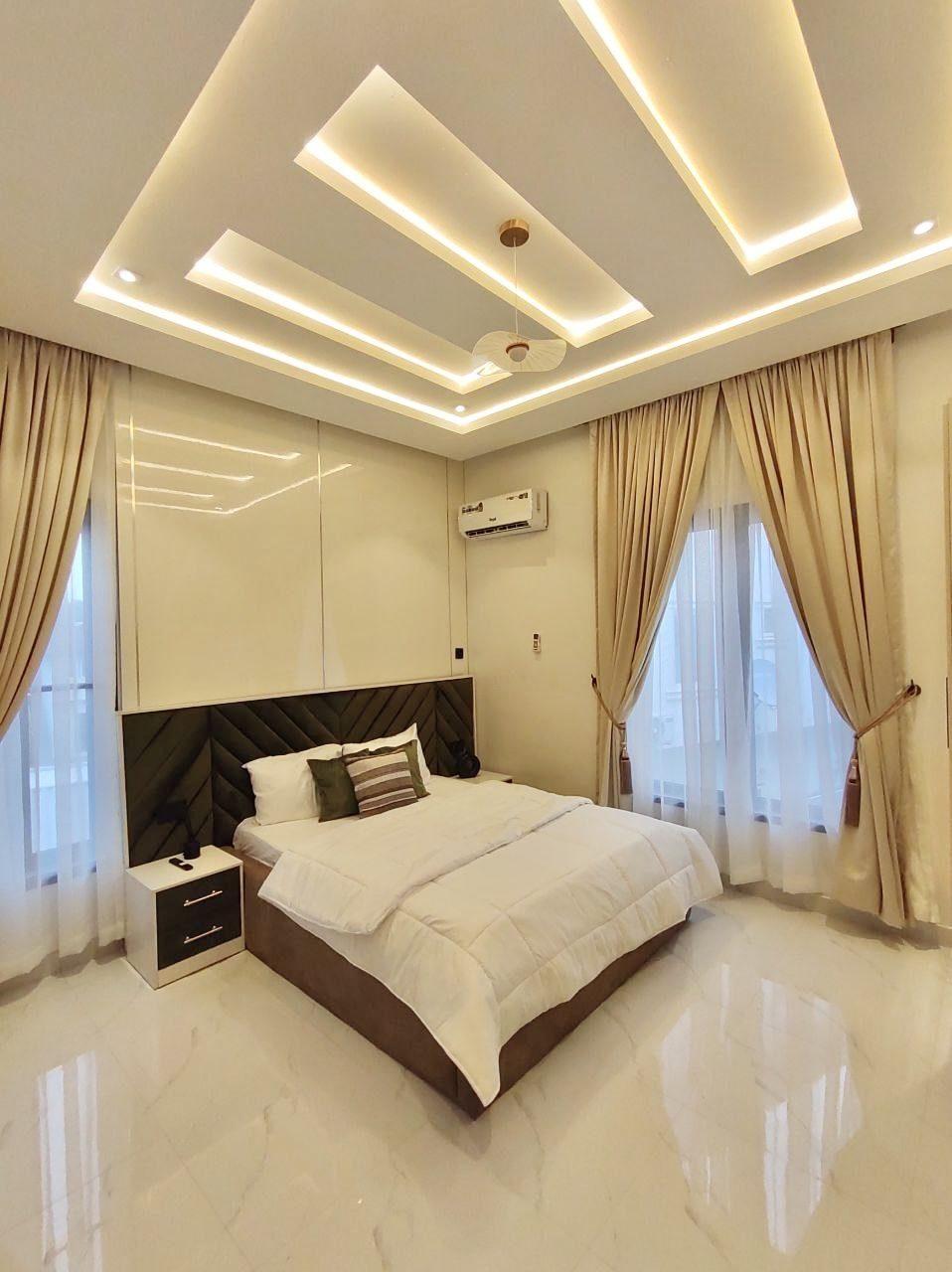 Bedroom with a layered lit ceiling, chevron upholstered headboard and full-height curtains