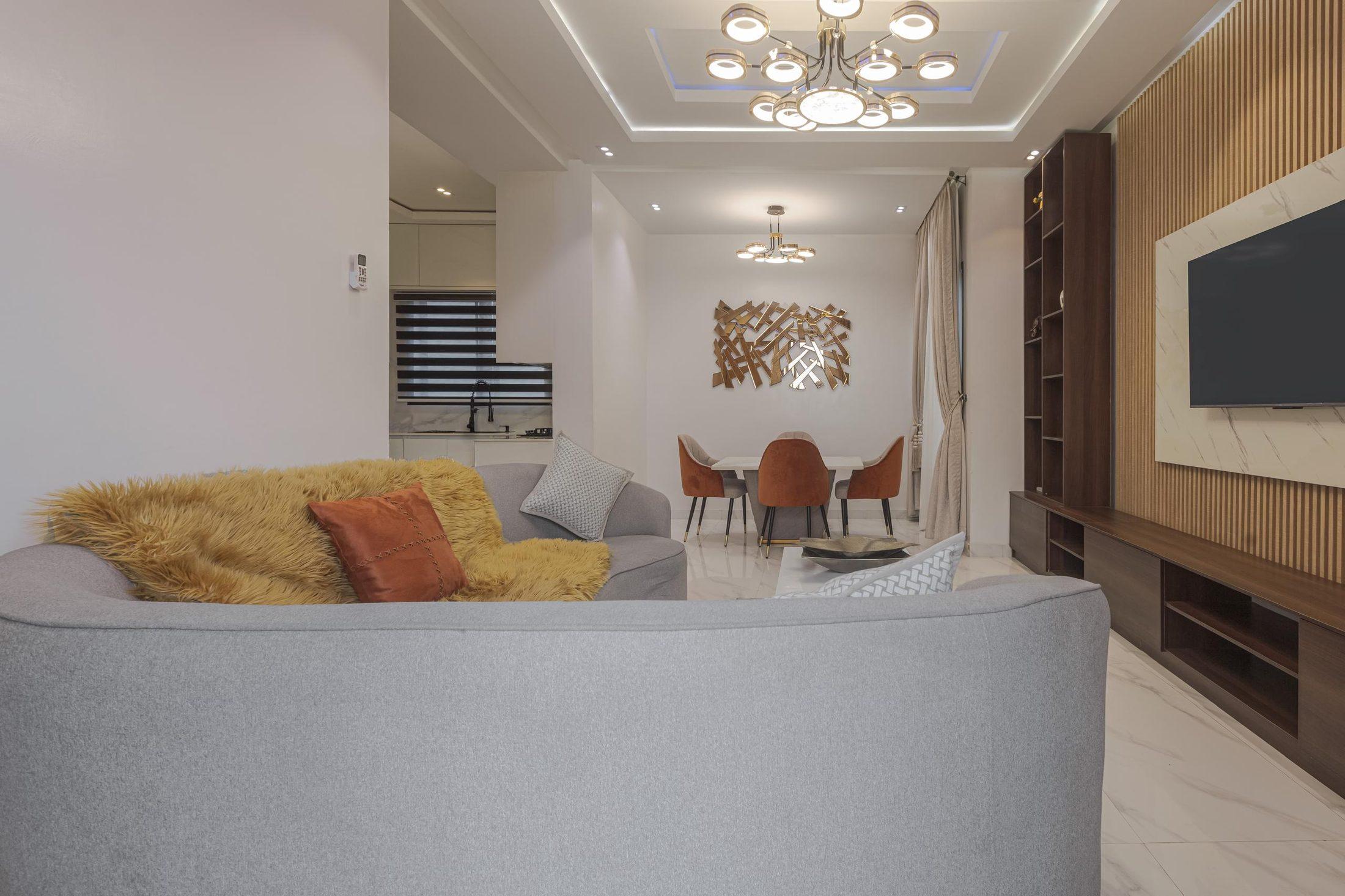 Open-plan living room with a soft grey curved sofa, timber-slatted media wall and a dining area beyond under a sculptural chandelier